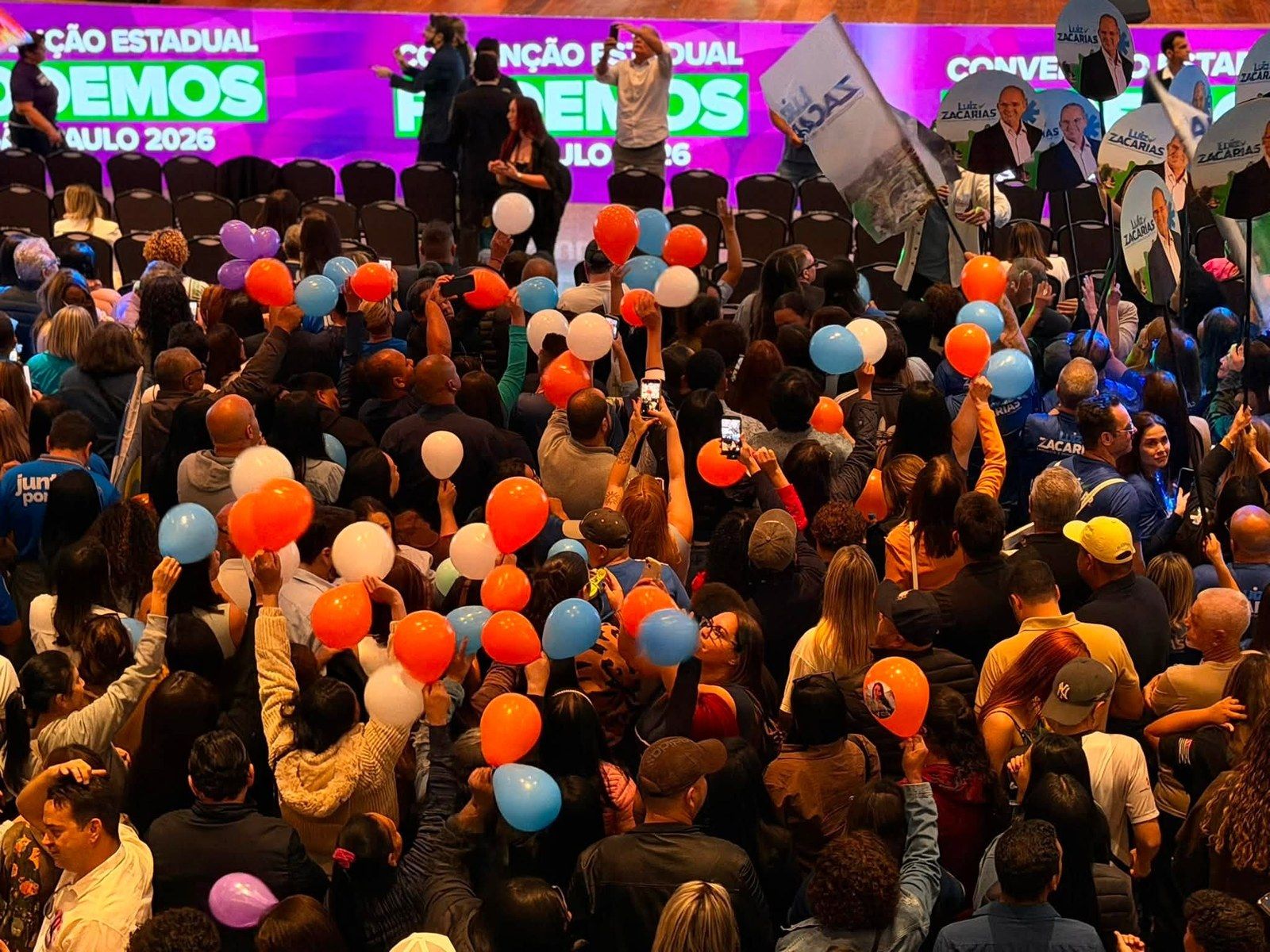 Multidão reunida diante de um palco durante uma convenção estadual, com balões e cartazes erguidos.