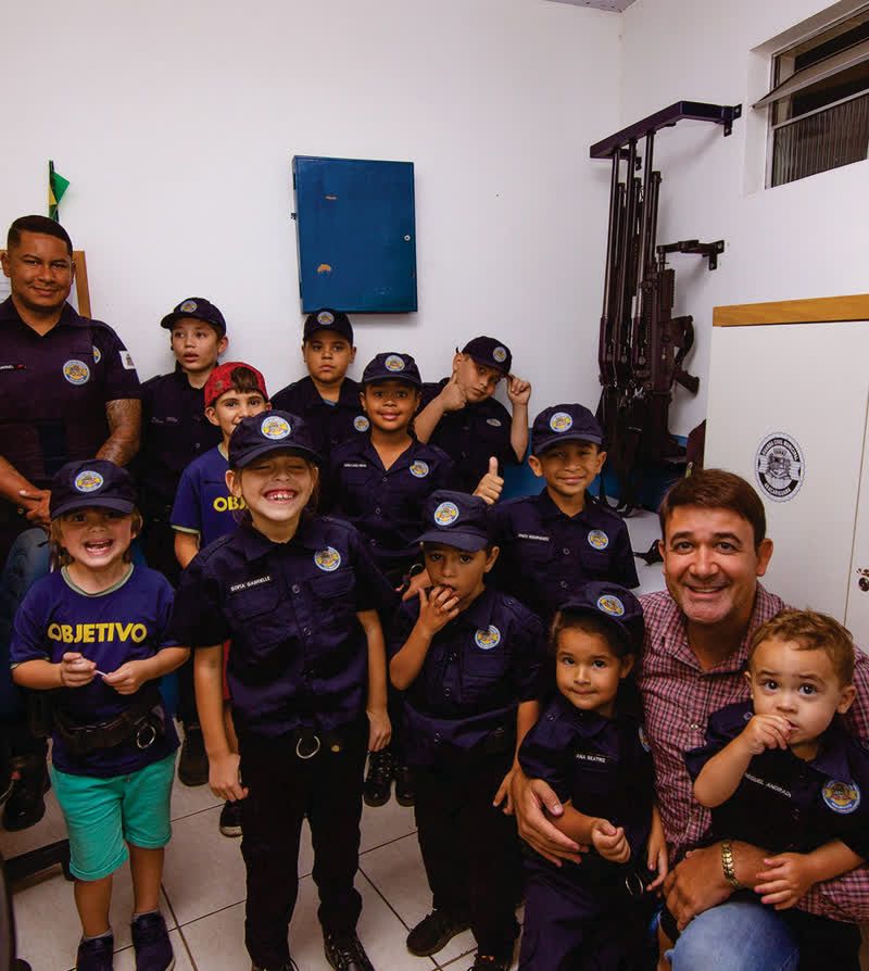 Crianças uniformizadas do Projeto Guarda Mirim posam ao lado de adultos.